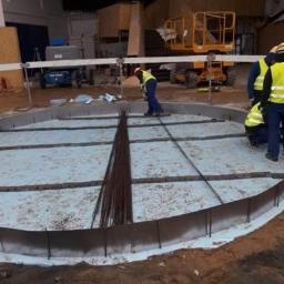 WM BUDSTAR Sp.z o.o . Sp.k. - Okrągły fundament w trakcie zbrojenia: widoczne pręty zbrojeniowe, szalunek z metalu i pracownicy w odblaskowych kamizelkach, przygotowujący podłoże na izolacji termicznej w hali budowlanej.