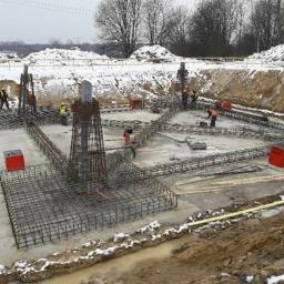 WM BUDSTAR Sp.z o.o . Sp.k. - Przygotowanie zbrojenia fundamentów pod budynek w zimowej scenerii; widoczne stalowe pręty tworzące szkielet, betonowa płyta fundamentowa, pracownicy w odblaskowych kamizelkach. W tle zasnieżony...