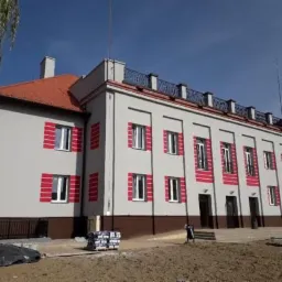 Odnowiony budynek z szarą elewacją, czerwonymi akcentami wokół okien i brązowym pasem u dołu, widoczne materiały budowlane na placu przed budynkiem.