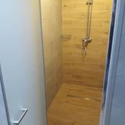 WM BUDSTAR Sp.z o.o . Sp.k. - Nowoczesna kabina prysznicowa z przeszklonymi drzwiami, wykończona płytkami imitującymi drewno na ścianach i podłodze, widoczny chromowany zestaw prysznicowy z deszczownicą i półką na kosmetyki.