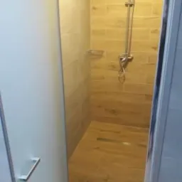 Nowoczesna kabina prysznicowa z przeszklonymi drzwiami, wykończona płytkami imitującymi drewno na ścianach i podłodze, widoczny chromowany zestaw prysznicowy z deszczownicą i półką na kosmetyki.