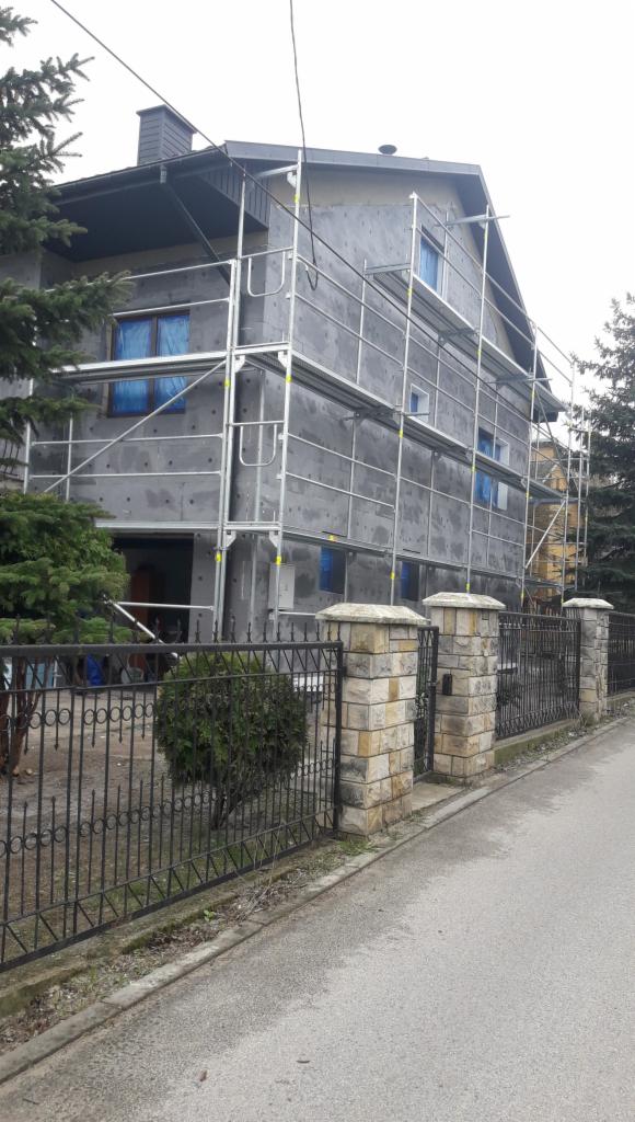 Dom jednorodzinny w trakcie prac elewacyjnych, widoczne rusztowania i zabezpieczone okna folią, ściany pokryte szarym materiałem izolacyjnym z widocznymi punktami mocowania.