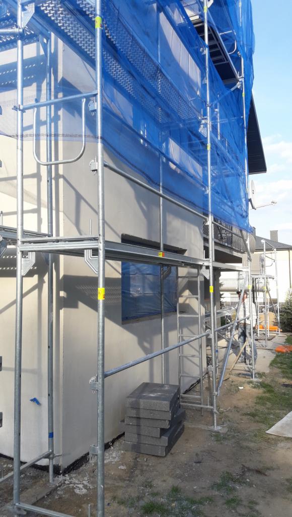 Ocieplanie elewacji domu jednorodzinnego z użyciem rusztowania i siatki ochronnej, widoczne płyty izolacyjne czekające na montaż.