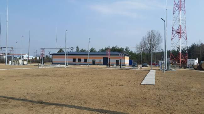 Budynek stacji transformatorowej z pomalowaną elewacją w biało-pomarańczowe pasy, widoczna wieża telekomunikacyjna w tle, teren przygotowany pod inwestycję.