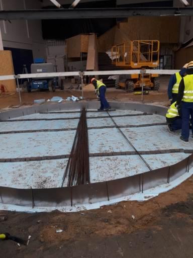 Okrągły fundament w trakcie zbrojenia: widoczne pręty zbrojeniowe, szalunek z metalu i pracownicy w odblaskowych kamizelkach, przygotowujący podłoże na izolacji termicznej w hali budowlanej.