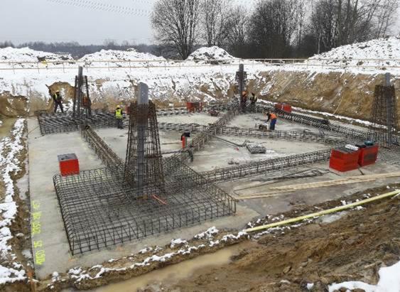 Przygotowanie zbrojenia fundamentów pod budynek w zimowej scenerii; widoczne stalowe pręty tworzące szkielet, betonowa płyta fundamentowa, pracownicy w odblaskowych kamizelkach. W tle zasnieżony...