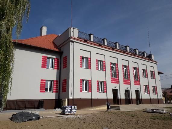 Odnowiony budynek z szarą elewacją, czerwonymi akcentami wokół okien i brązowym pasem u dołu, widoczne materiały budowlane na placu przed budynkiem.