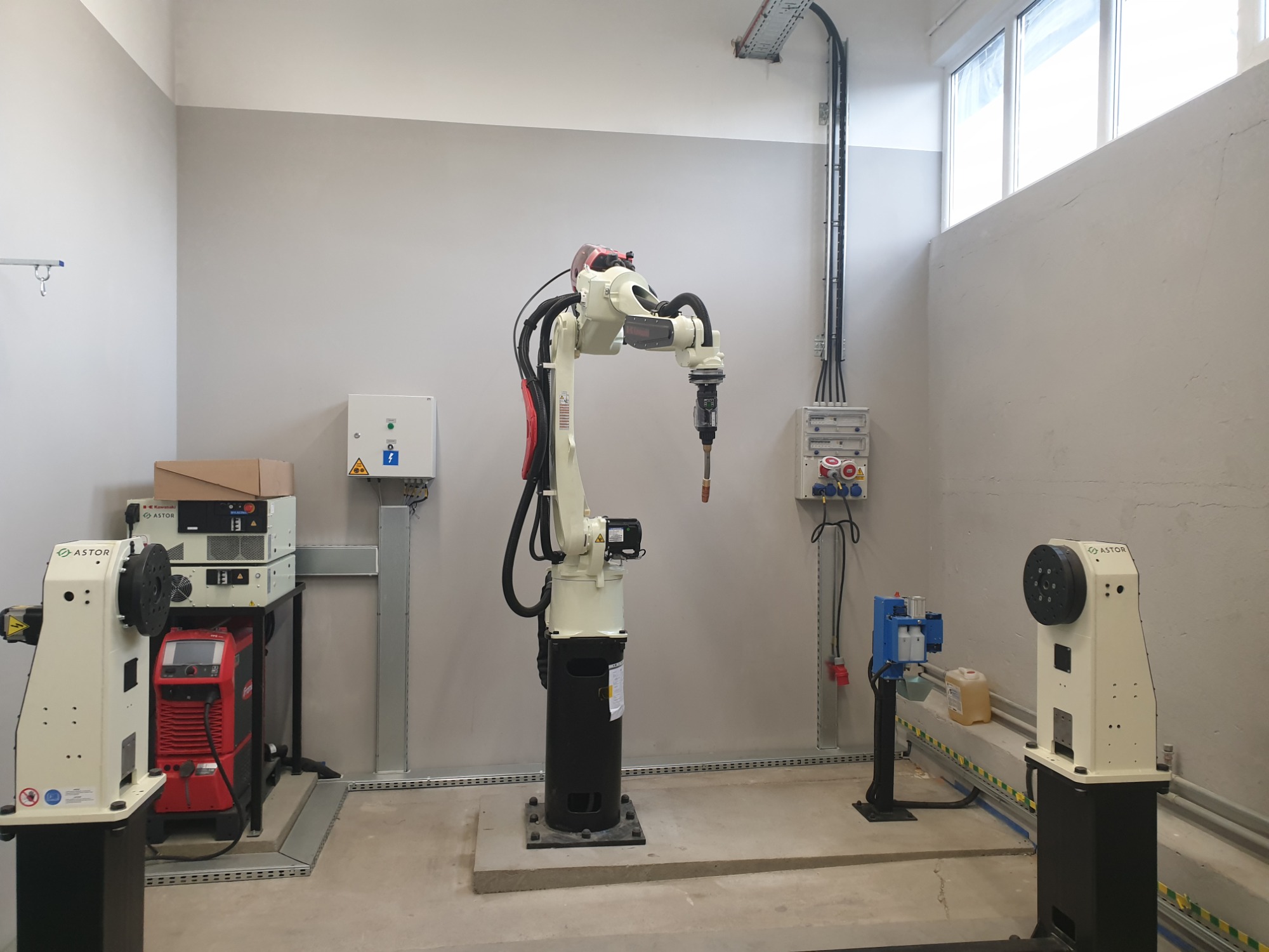 Wnętrze hali produkcyjnej z robotem spawalniczym Kawasaki w centrum, obok spawarka Fronius i osprzęt Astor, na ścianie skrzynki elektryczne i kable w korytach.