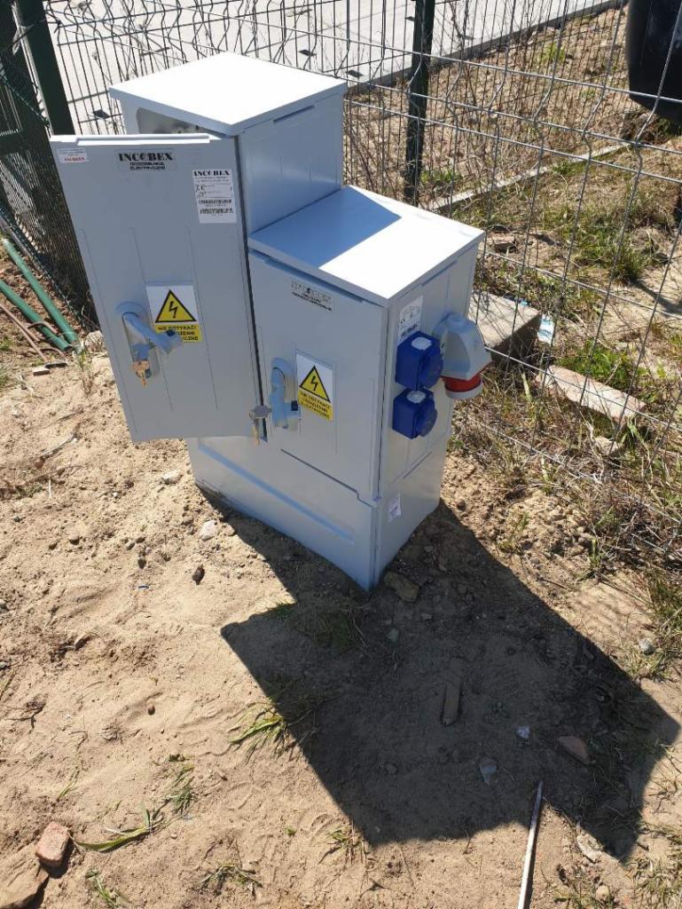 Szara, metalowa skrzynka elektryczna z otwartymi drzwiczkami i gniazdami na zewnątrz, umieszczona na piaszczystym podłożu z widocznym ogrodzeniem z siatki w tle.