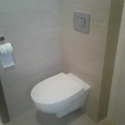 Nowoczesna toaleta podwieszana z szarymi płytkami imitującymi drewno, srebrny przycisk spłukiwania i akcesoria łazienkowe na półce.