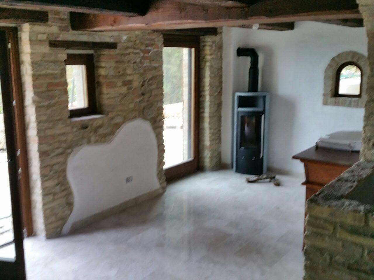 Wnętrze pokoju z surową cegłą, przygotowane pod gładzie, z widocznym kominkiem wolnostojącym i drewnianymi belkami stropowymi.