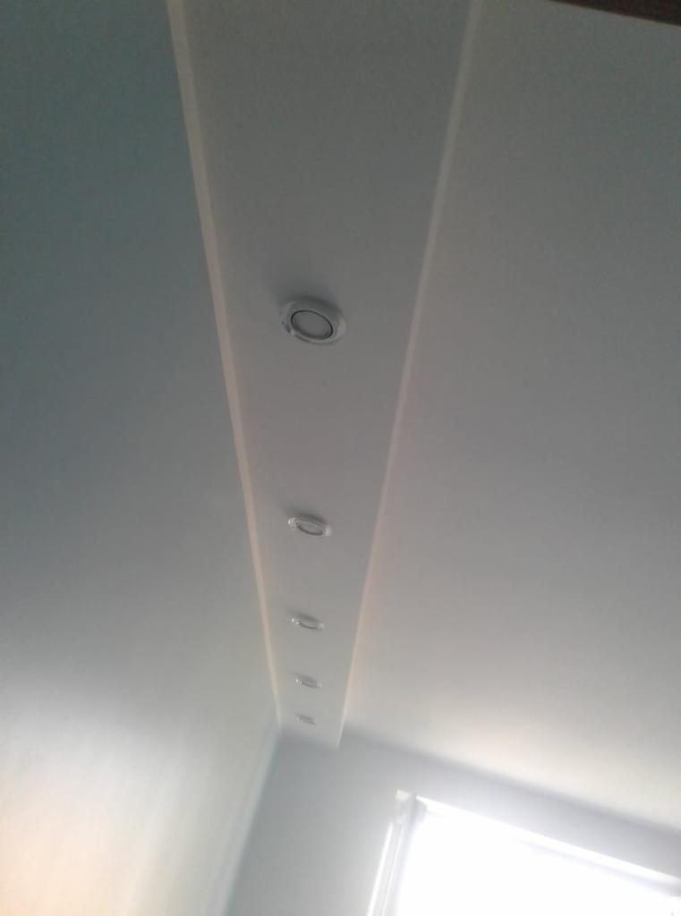 Biały sufit podwieszany z zamontowanymi okrągłymi lampami halogenowymi, widoczne światło dzienne z okna w rogu.