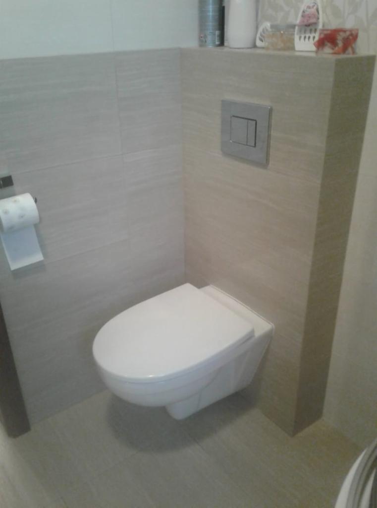 Nowoczesna toaleta podwieszana z szarymi płytkami imitującymi drewno, srebrny przycisk spłukiwania i akcesoria łazienkowe na półce.