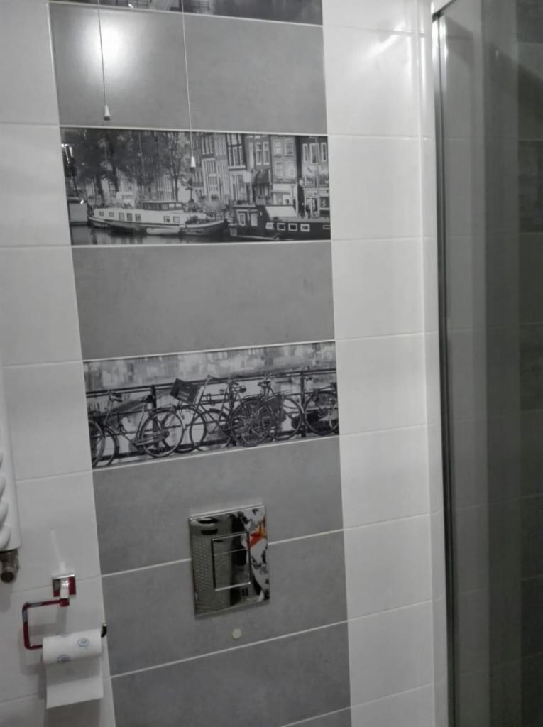 Wykończona łazienka z szarymi i białymi płytkami z motywami czarno-białych zdjęć holenderskich krajobrazów i rowerów, widoczny fragment kabiny prysznicowej i uchwyt na papier toaletowy.