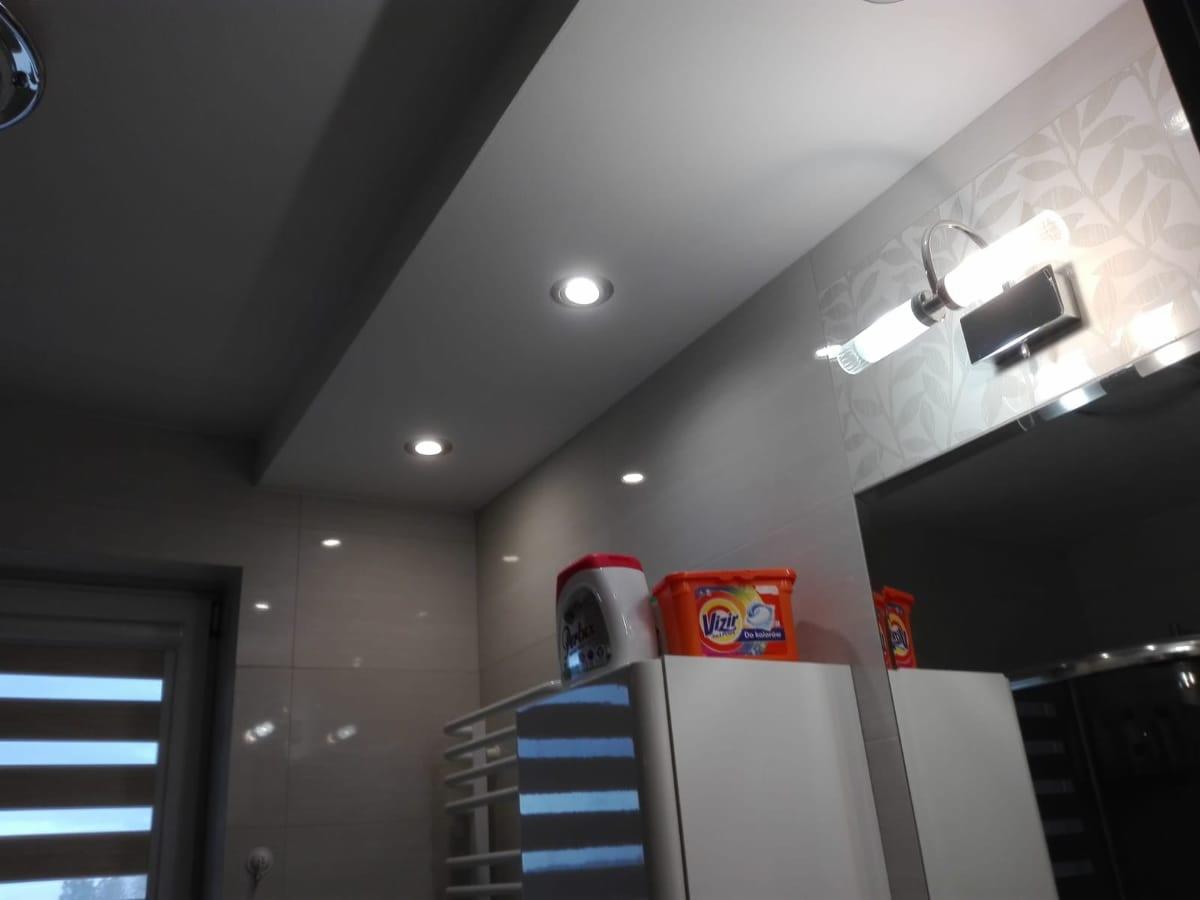 Wykończona łazienka z podwieszanym sufitem z oświetleniem punktowym, lustrem z podświetleniem, białą szafką i grzejnikiem drabinkowym. Na szafce widoczne pojemniki z detergentami.