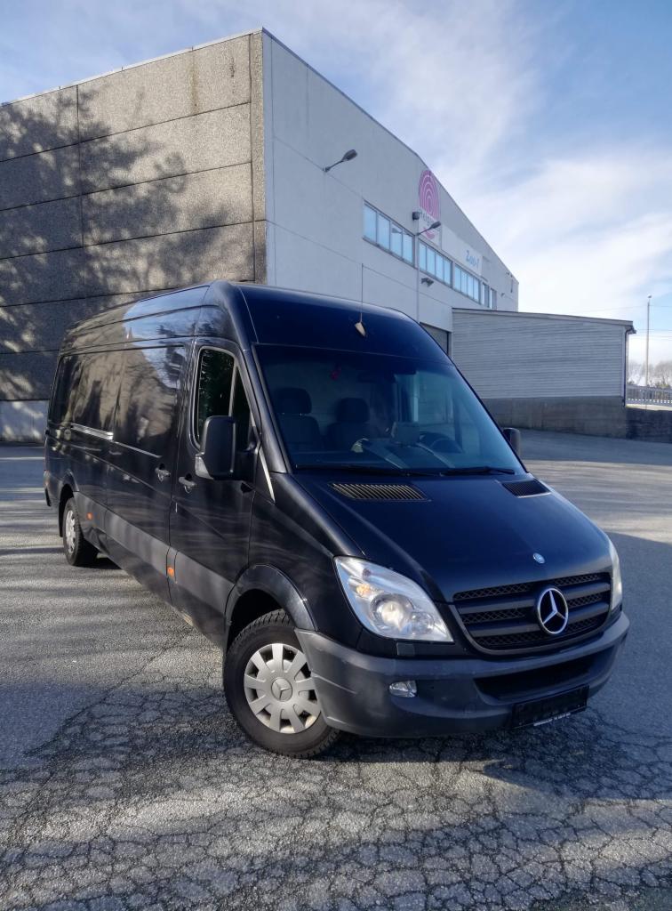 Czarny bus Mercedes-Benz Sprinter firmy transportowej z Augustowa, zaparkowany przed budynkiem magazynowym w słoneczny dzień.