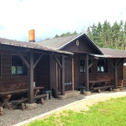 Ośrodek Szkolenia Sportowego Brzednia - Drewniany budynek z numerem 6 na dachu, z zadaszonymi werandami, ławami i stołami na zewnątrz, otoczony drzewami i trawnikiem.