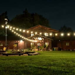 Ośrodek Szkolenia Sportowego Brzednia - Oświetlony drewniany budynek z zielonym trawnikiem, girlandami świetlnymi i miejscem na ognisko z ławkami, wieczorową porą.