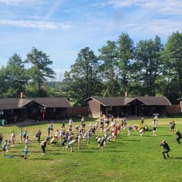 Ośrodek Szkolenia Sportowego Brzednia - Grupa dzieci i dorosłych ćwiczy na zielonej trawie przed drewnianymi domkami letniskowymi w słoneczny dzień, widok z góry.
