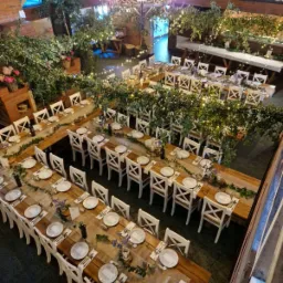 Uroczysta sala bankietowa z drewnianymi stołami, białymi krzesłami, dekoracjami z zieleni i subtelnym oświetleniem, przygotowana na przyjęcie z widokiem z góry.