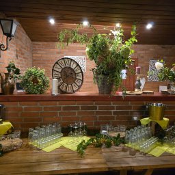 Ośrodek Szkolenia Sportowego Brzednia - Rustykalna aranżacja stołu z dużą ilością szklanek, dekoracjami kwiatowymi i zegarem na tle ceglanej ściany, oświetlona punktowymi lampami.