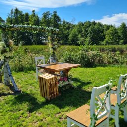 Ośrodek Szkolenia Sportowego Brzednia - Uroczysta aranżacja plenerowa z drewnianym stołem, białymi krzesłami zdobionymi bluszczem i konstrukcją z brzozowych pni udekorowaną kwiatami, na tle zieleni i błękitnego nieba.