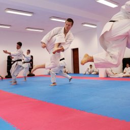 Ośrodek Szkolenia Sportowego Brzednia - Grupa osób w białych strojach do karate ćwiczy kopnięcia w sali z czerwono-niebieską matą podłogową, z widocznymi workami treningowymi i siedzącymi osobami w tle.