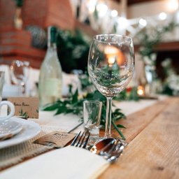 Ośrodek Szkolenia Sportowego Brzednia - Elegancko nakryty stół z drewnianym blatem, kieliszkiem do wina, szklanką, sztućcami, białą zastawą i zielonymi dekoracjami, w tle ceglany kominek i girlanda z lampek.