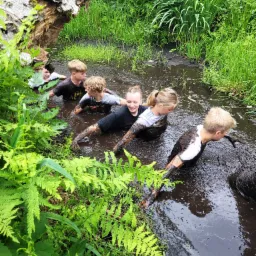 Grupa dzieci brnących przez błotniste, zarośnięte bagnisko podczas zabawy na świeżym powietrzu, z powalonym drzewem w tle.