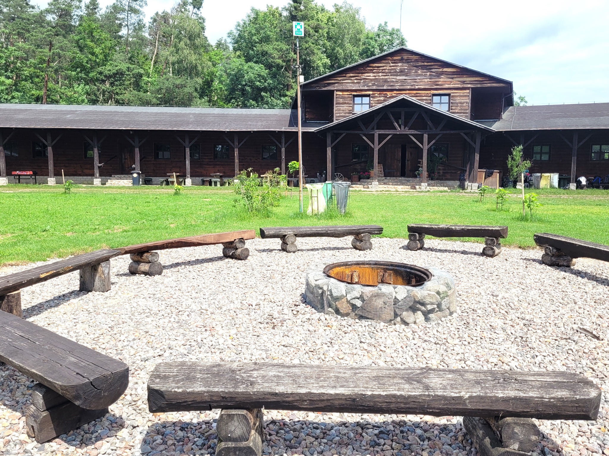 Ognisko otoczone drewnianymi ławkami na terenie ośrodka wypoczynkowego z drewnianym budynkiem w tle, otoczone zielenią.