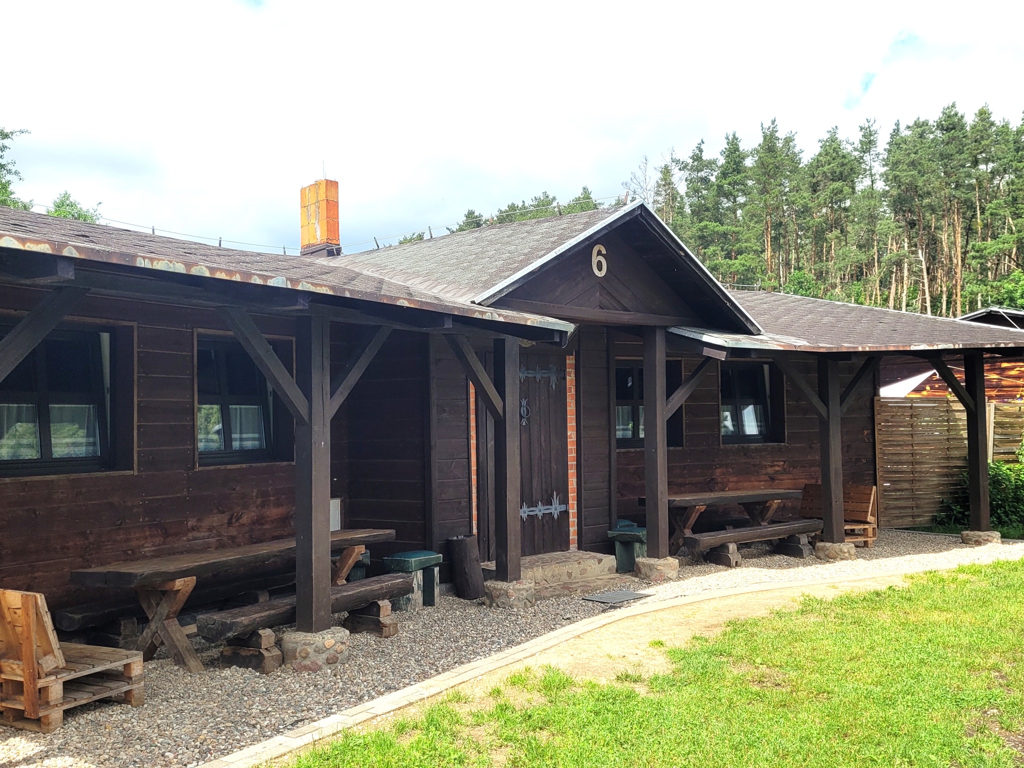 Drewniany budynek z numerem 6 na dachu, z zadaszonymi werandami, ławami i stołami na zewnątrz, otoczony drzewami i trawnikiem.
