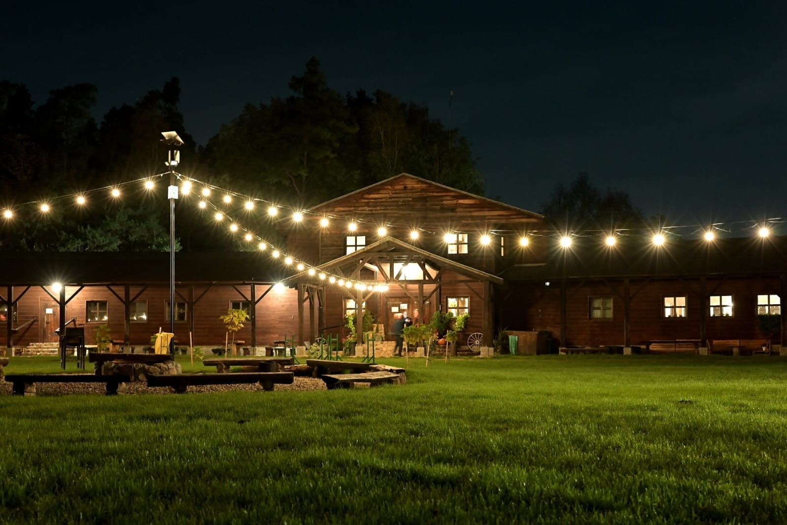 Oświetlony drewniany budynek z zielonym trawnikiem, girlandami świetlnymi i miejscem na ognisko z ławkami, wieczorową porą.