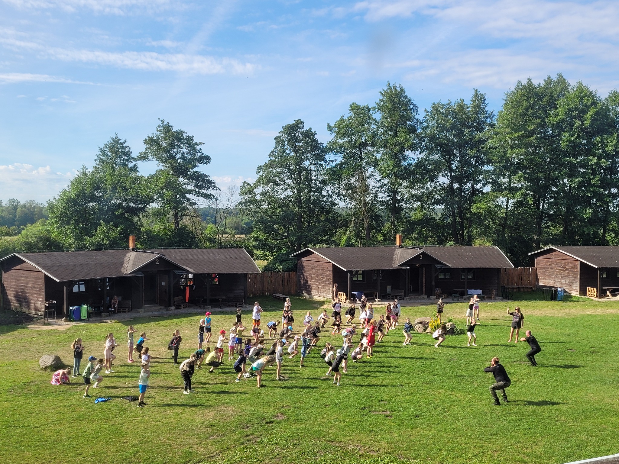Grupa dzieci i dorosłych ćwiczy na zielonej trawie przed drewnianymi domkami letniskowymi w słoneczny dzień, widok z góry.