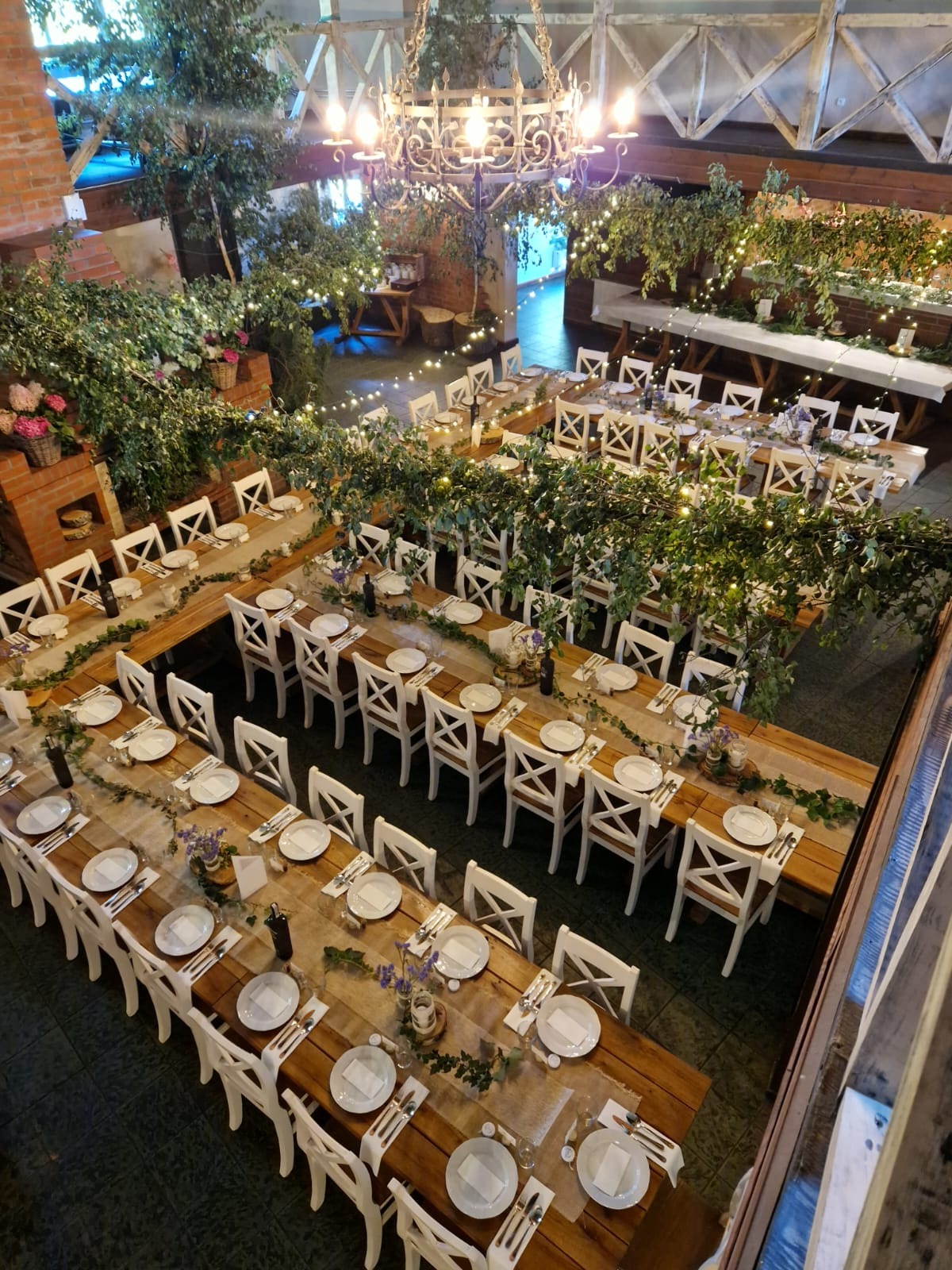 Uroczysta sala bankietowa z drewnianymi stołami, białymi krzesłami, dekoracjami z zieleni i subtelnym oświetleniem, przygotowana na przyjęcie z widokiem z góry.