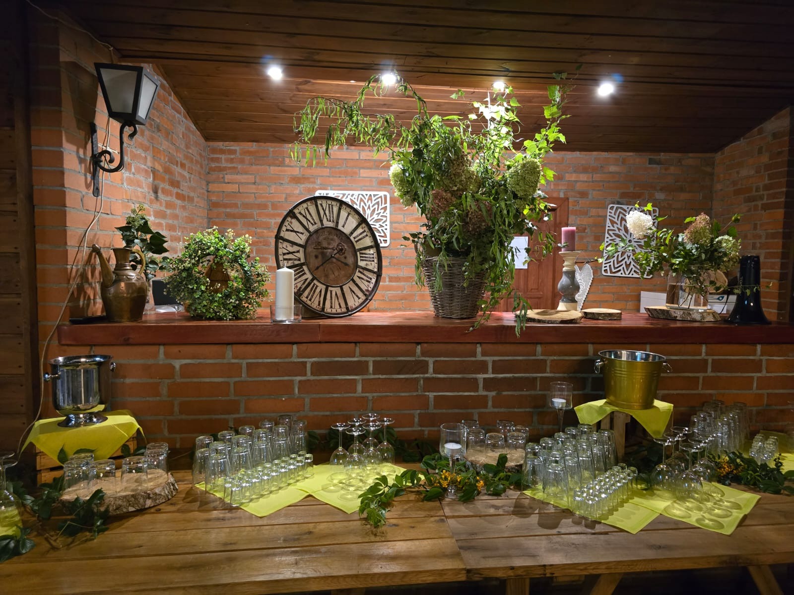 Rustykalna aranżacja stołu z dużą ilością szklanek, dekoracjami kwiatowymi i zegarem na tle ceglanej ściany, oświetlona punktowymi lampami.