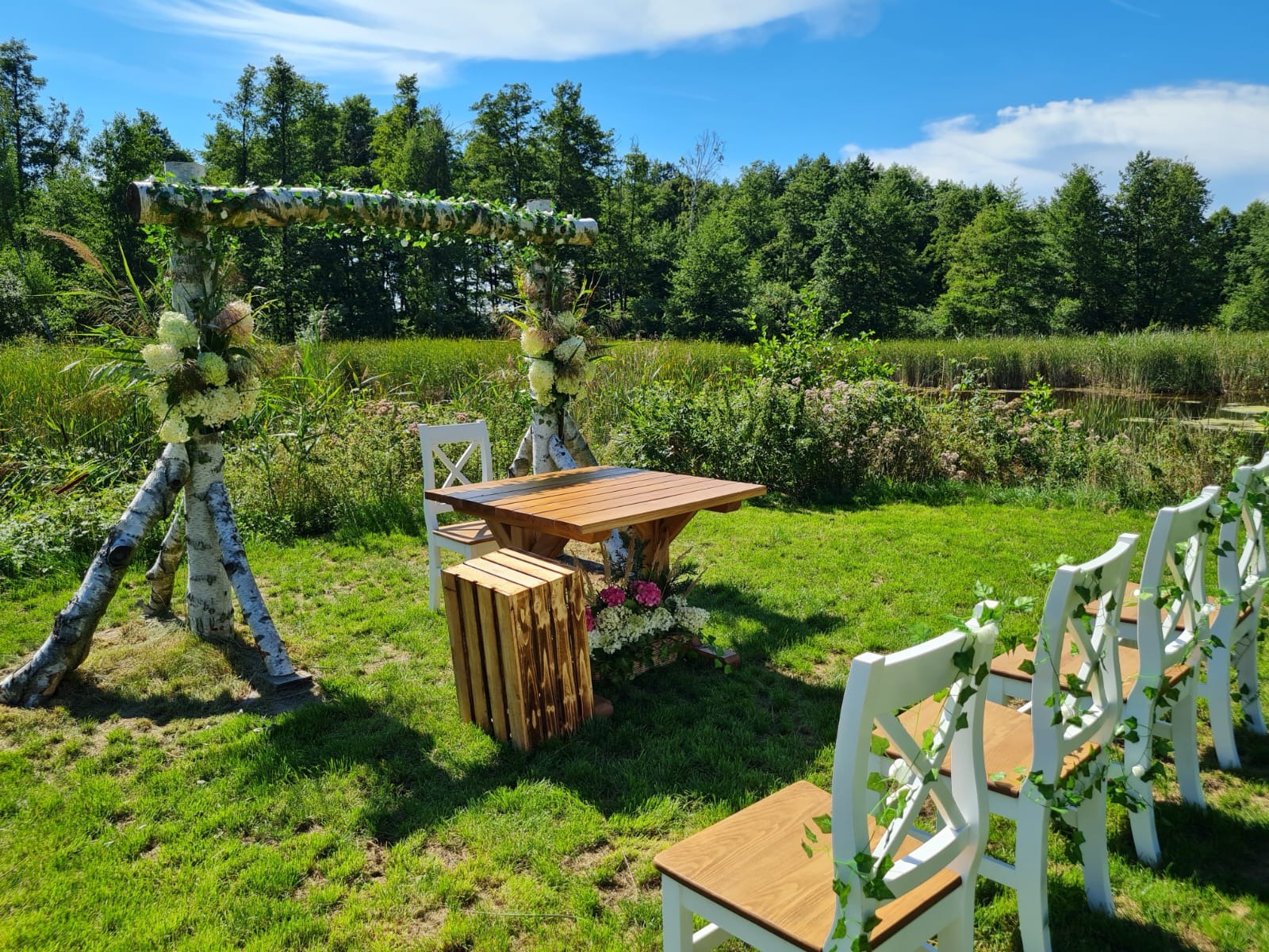 Uroczysta aranżacja plenerowa z drewnianym stołem, białymi krzesłami zdobionymi bluszczem i konstrukcją z brzozowych pni udekorowaną kwiatami, na tle zieleni i błękitnego nieba.