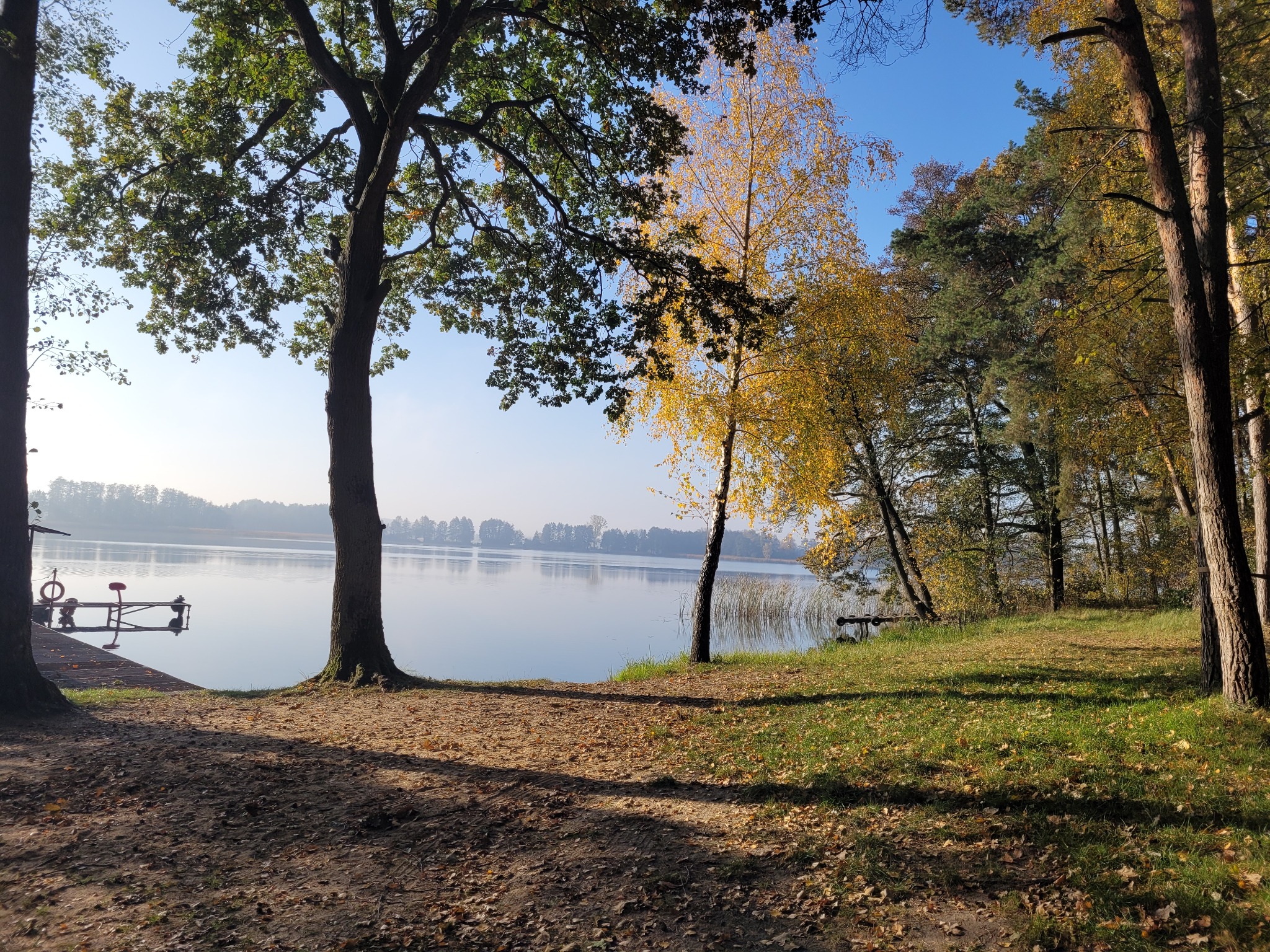 Spokojny krajobraz jeziora w jesiennej scenerii z pomostem, drzewami o barwnych liściach i mglistym horyzontem.