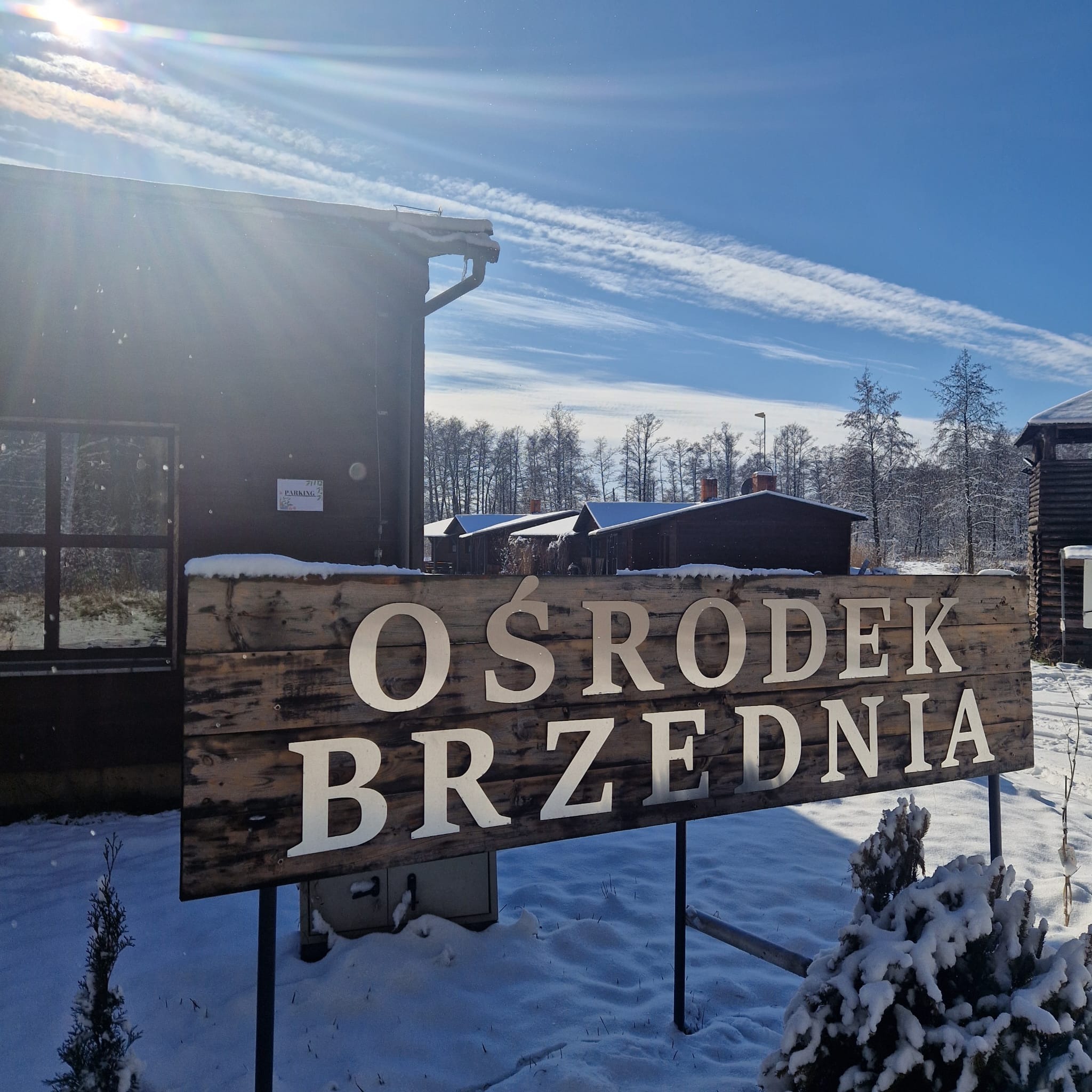 Tablica z napisem 'Ośrodek Brzednia' przed budynkami ośrodka w zimowej scenerii, pokryta śniegiem.