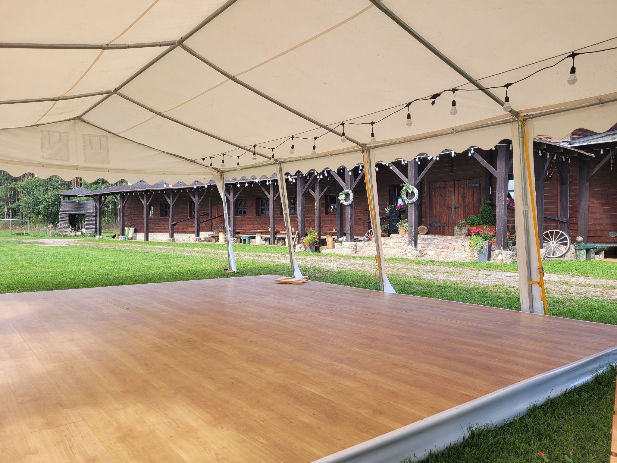 Drewniany parkiet taneczny pod namiotem eventowym, oświetlony girlandą żarówek, z widokiem na rustykalną drewnianą zabudowę i zielony trawnik.