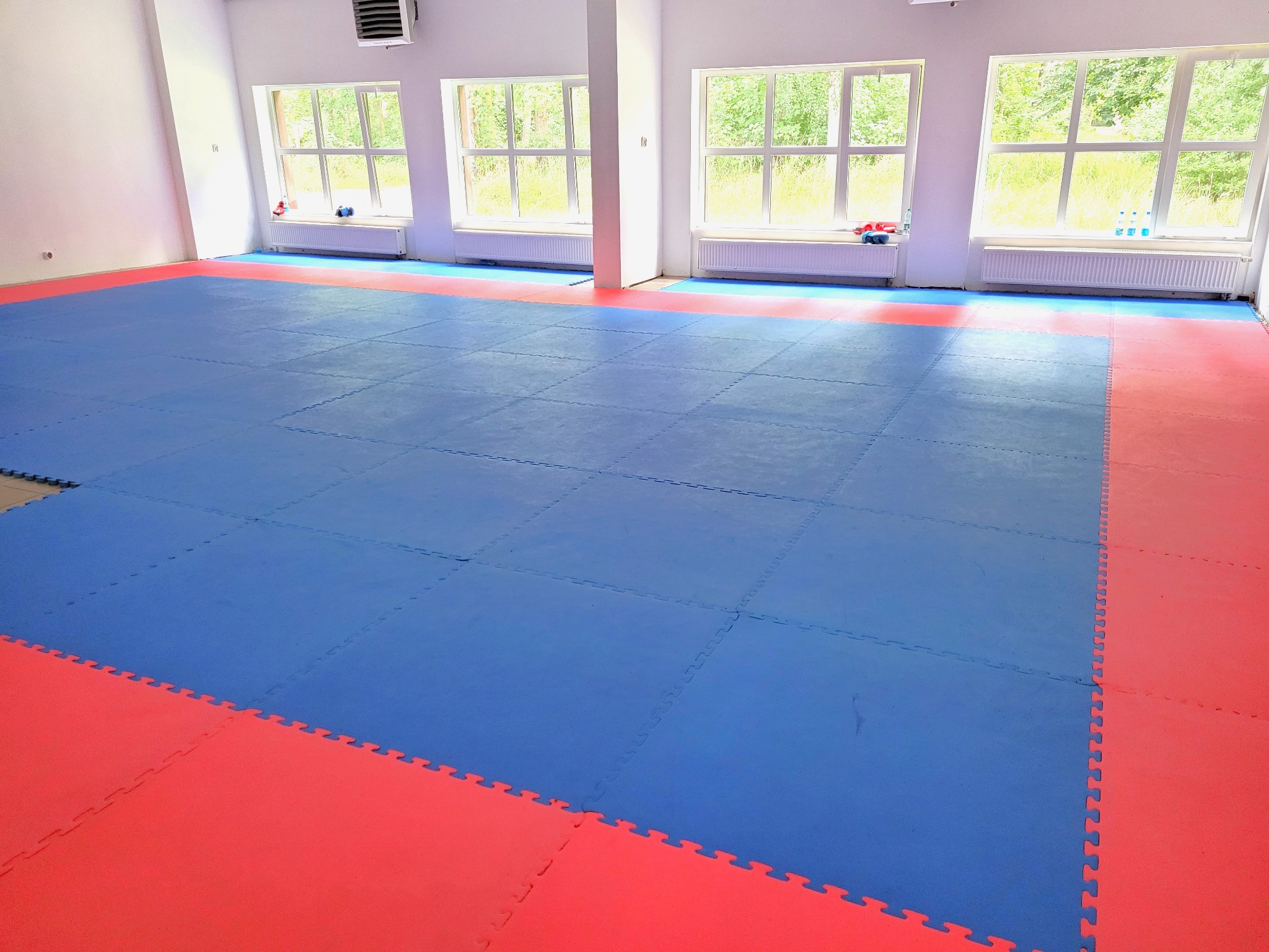 Pusta sala gimnastyczna z niebiesko-czerwonymi matami podłogowymi, jasne wnętrze z trzema oknami i grzejnikami, widok na zieleń za oknami.