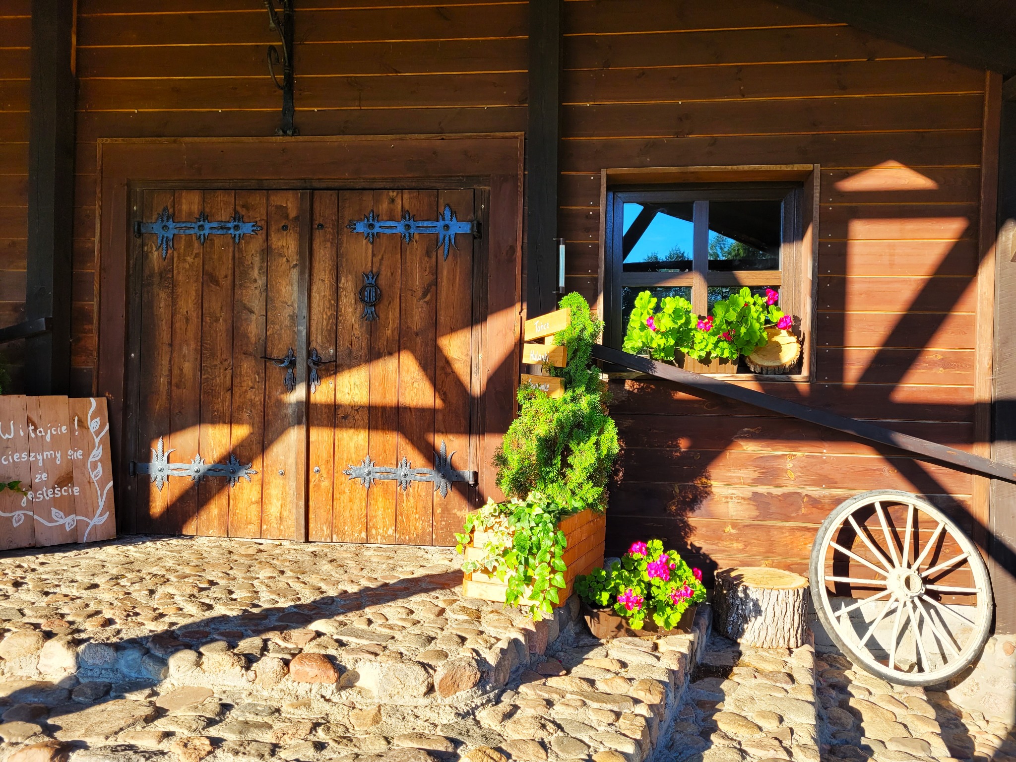 Rustykalne wejście do budynku z drewnianymi drzwiami z ozdobnymi zawiasami, okno z kwiatami, dekoracje roślinne, koło od wozu i tablica powitalna na kamiennych schodach.