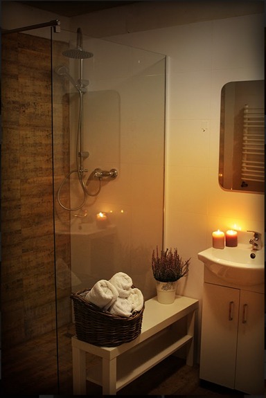 Nowoczesna łazienka z kabiną prysznicową typu walk-in, wiklinowym koszem z białymi ręcznikami, umywalką z szafką, świecami i doniczką z lawendą, oświetlona ciepłym światłem.