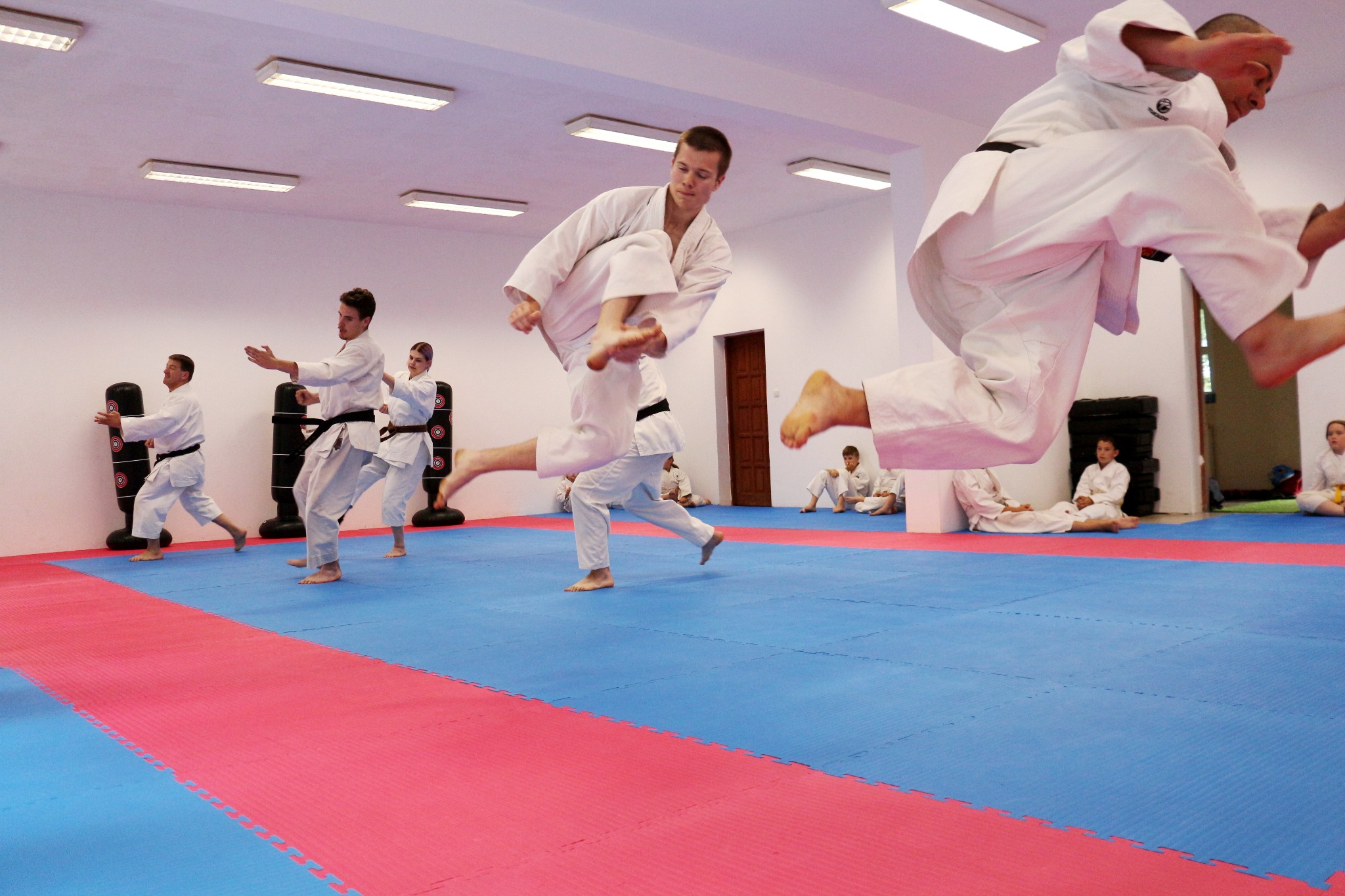 Grupa osób w białych strojach do karate ćwiczy kopnięcia w sali z czerwono-niebieską matą podłogową, z widocznymi workami treningowymi i siedzącymi osobami w tle.