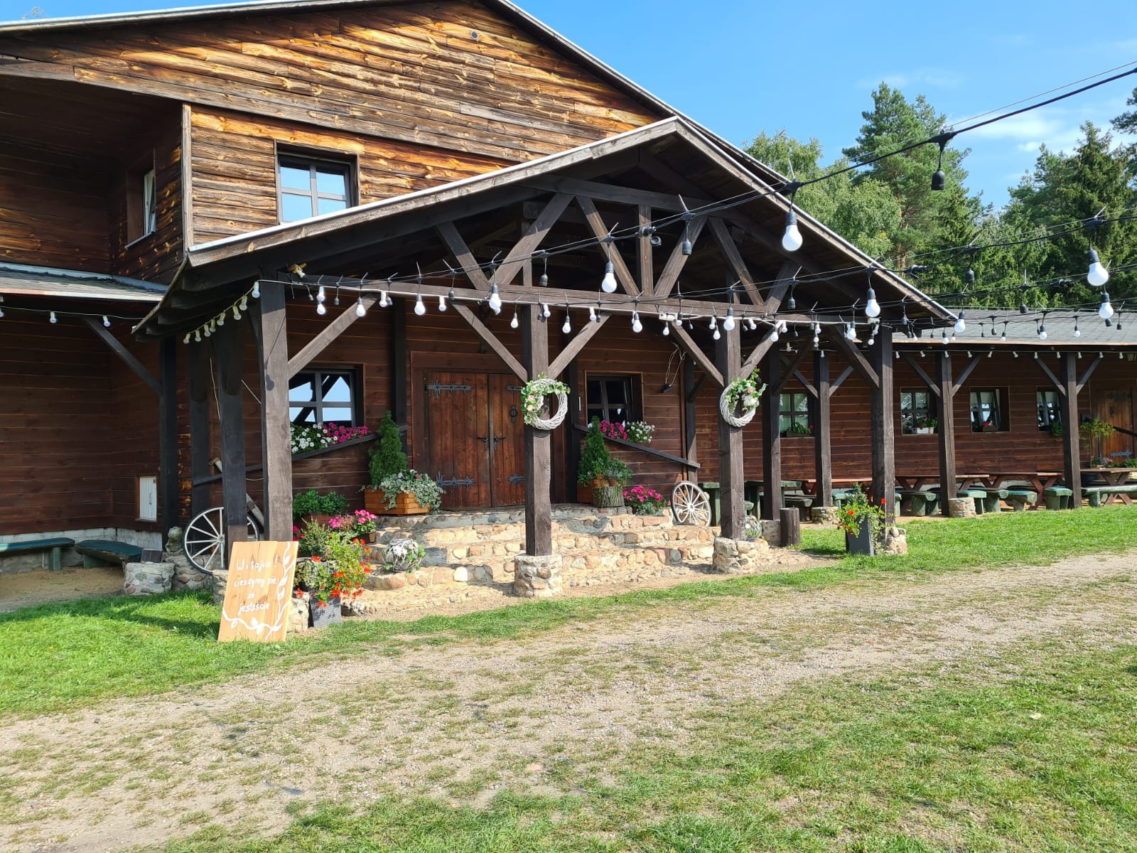 Rustykalna drewniana chata z werandą, dekoracjami kwiatowymi i oświetleniem girlandami żarówek, przygotowana na imprezę plenerową.