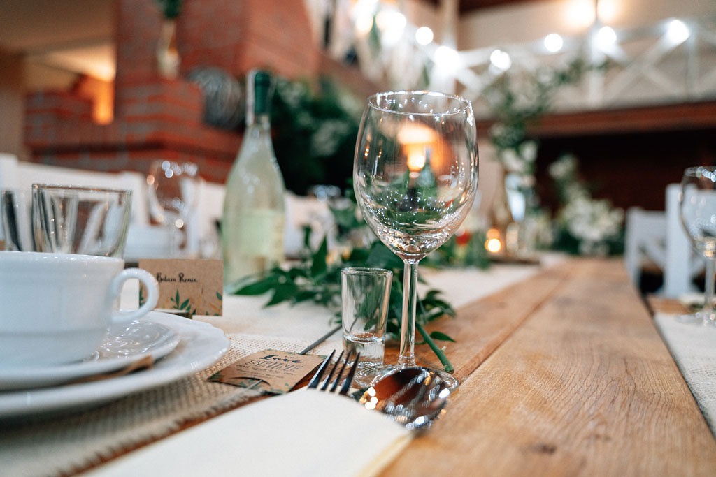 Elegancko nakryty stół z drewnianym blatem, kieliszkiem do wina, szklanką, sztućcami, białą zastawą i zielonymi dekoracjami, w tle ceglany kominek i girlanda z lampek.