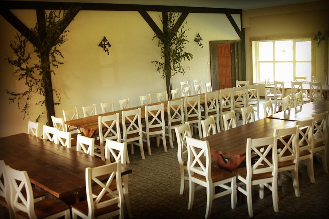 Sala bankietowa z długimi drewnianymi stołami i białymi krzesłami typu crossback, dekorowana malowanymi drzewami na ścianach i metalowymi kinkietami.