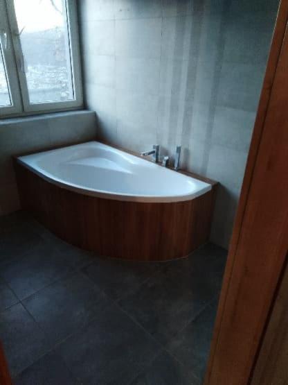 Nowoczesna, narożna wanna obudowana drewnopodobną okładziną, z baterią umywalkową, na tle szarych płytek ściennych i podłogowych, z oknem w tle.