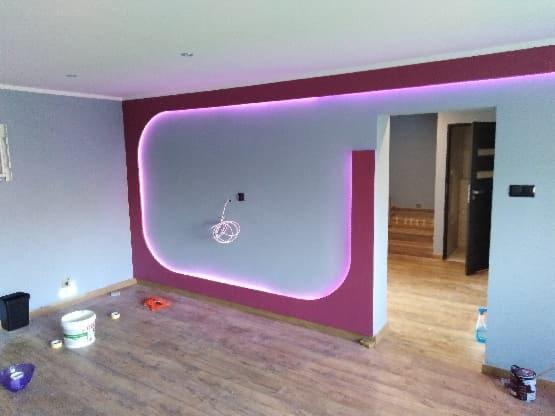 Nowoczesne wnętrze po remoncie: ściana pomalowana na szaro z bordową obwódką podświetloną fioletowym światłem LED, widoczne narzędzia malarskie i wiązka kabli wystająca ze ściany.