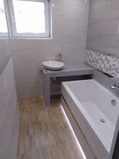 Nowoczesna łazienka z białymi płytkami imitującymi drewno na ścianach, podświetlaną wanną, umywalką nablatową na szarym blacie i drewnopodobną podłogą.