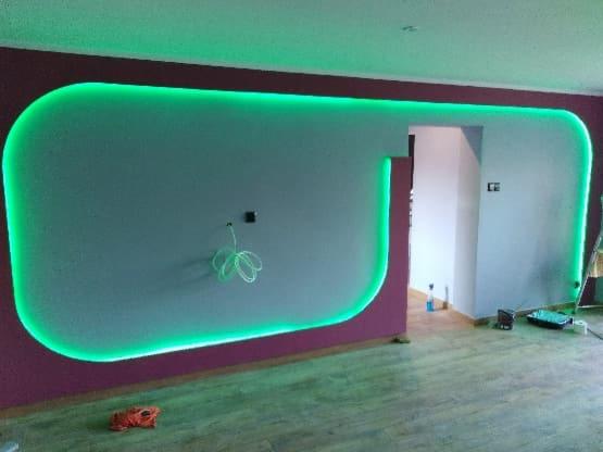 Ściana z wnęką oświetloną zielonymi ledami, widoczne przewody, podłoga z paneli, w tle otwarte przejście do innego pomieszczenia.